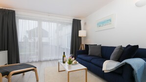 A modern living room with a dark blue sofa, a white coffee table, and a bench. There is a lamp, a picture on the wall, and a window with sheer curtains. - 21 Carthew Court - Garage Parking - Outside Space - Dog Friendly (St Ives)