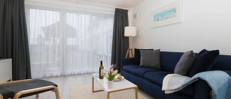 Un salon moderne avec un canapé bleu foncé, une table basse blanche et un banc. On y trouve une lampe, un tableau au mur et une fenêtre avec des rideaux légers.