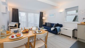 A modern living room with a sofa, a dining table with breakfast items, and a view of the outside through a window. - 21 Carthew Court - Garage Parking - Outside Space - Dog Friendly (St Ives)