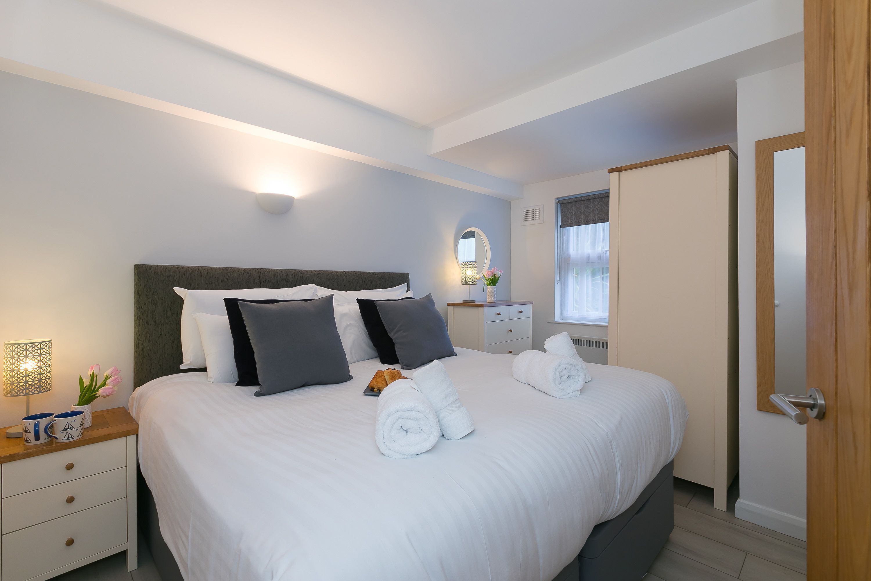 Un lit bien fait, avec du linge de lit blanc et des serviettes pliées, une table de chevet avec une lampe et un vase de fleurs, et une porte en bois sur la droite.