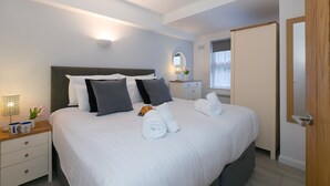 A neatly made bed with white linens and rolled towels, a bedside table with a lamp and a vase of flowers, and a wooden door to the right. - 21 Carthew Court - Garage Parking - Outside Space - Dog Friendly (St Ives)