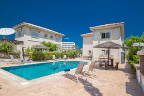 A swimming pool with lounge chairs and umbrellas, surrounded by buildings with tiled roofs and windows. - Jason 8  Villa (Paralimni)