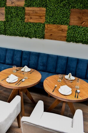 A dining area with two round wooden tables set for two, each with white napkins, cutlery, and glasses. The tables are placed on a wooden floor, and there is a blue upholstered bench in the background. - Tecno Boutique Hotel (La Paz)