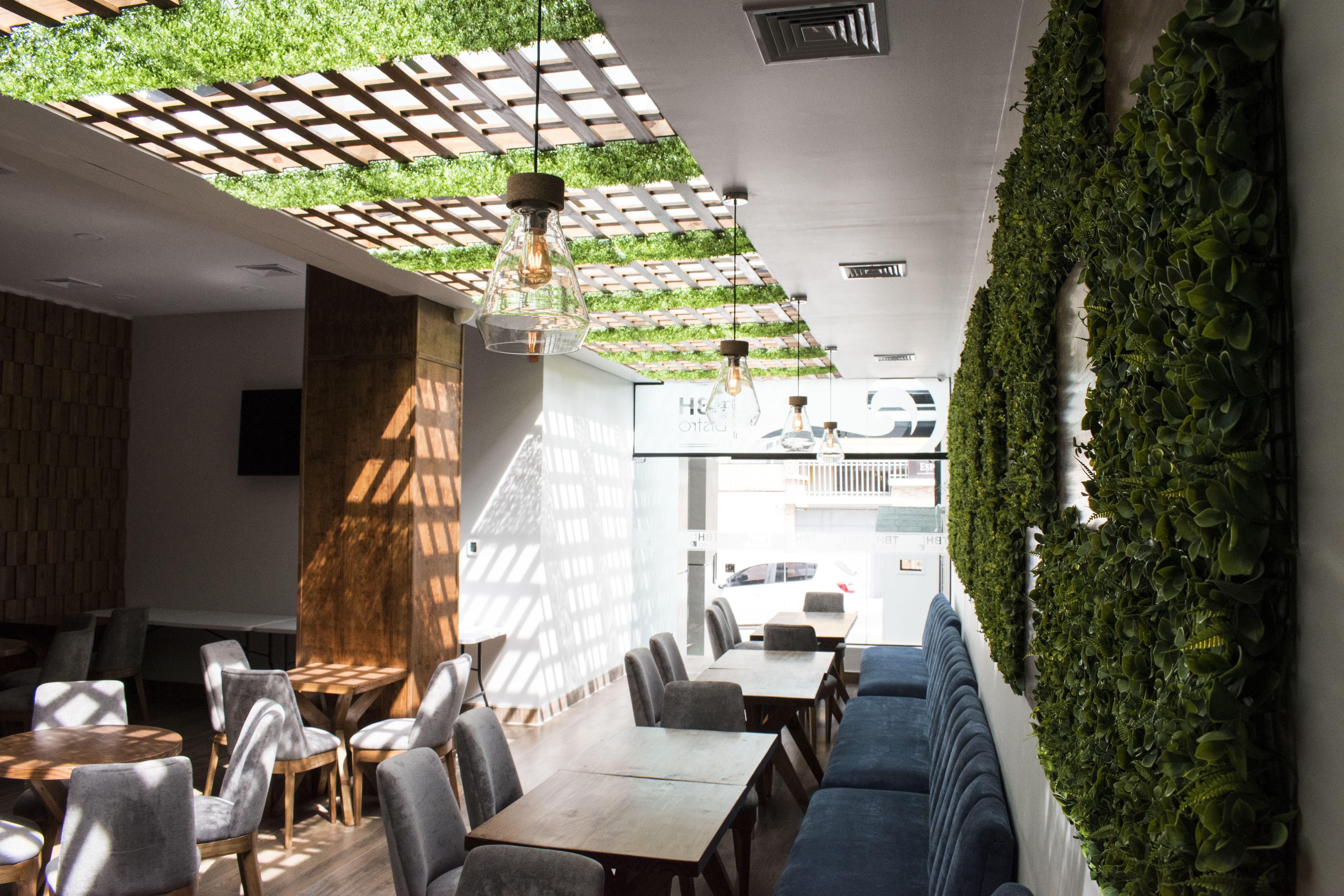 Un interior de restaurante moderno con mesas y sillas de madera, un sofá azul y una pared verde con plantas.
