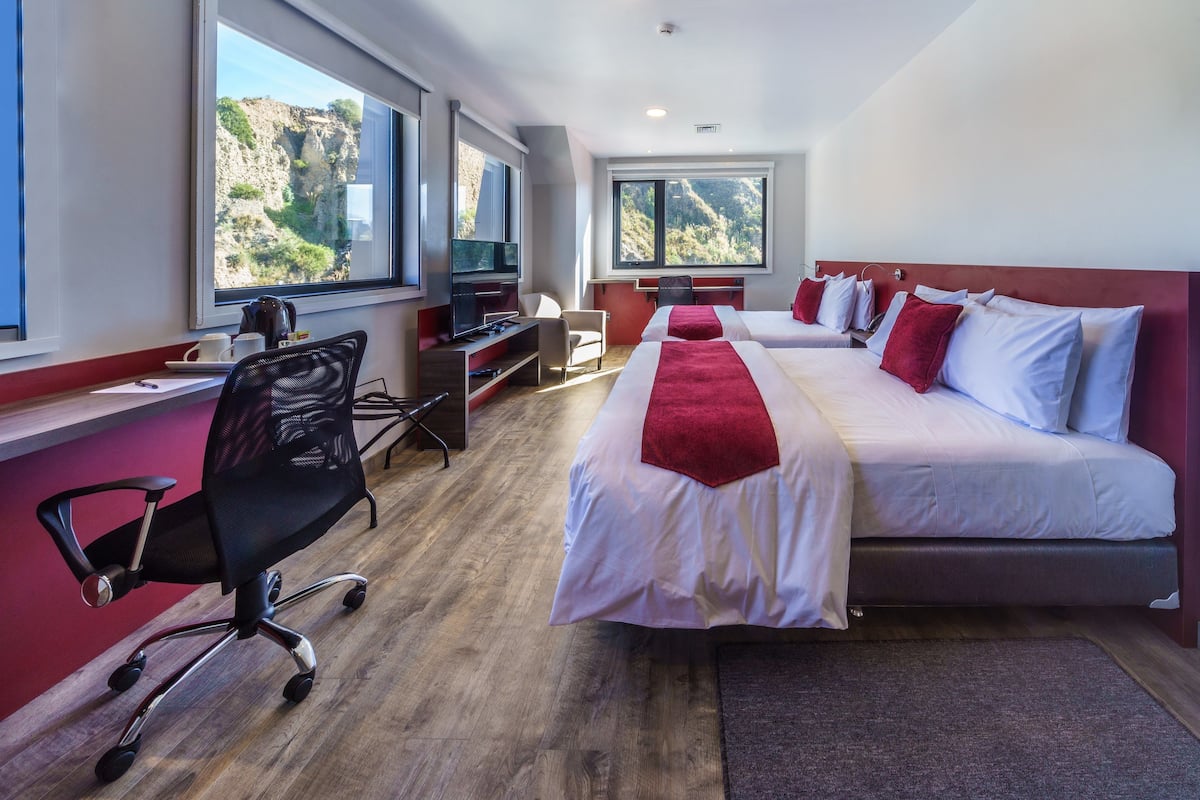 Une chambre d’hôtel avec deux lits, un bureau, une chaise et de grandes fenêtres donnant sur l’extérieur.