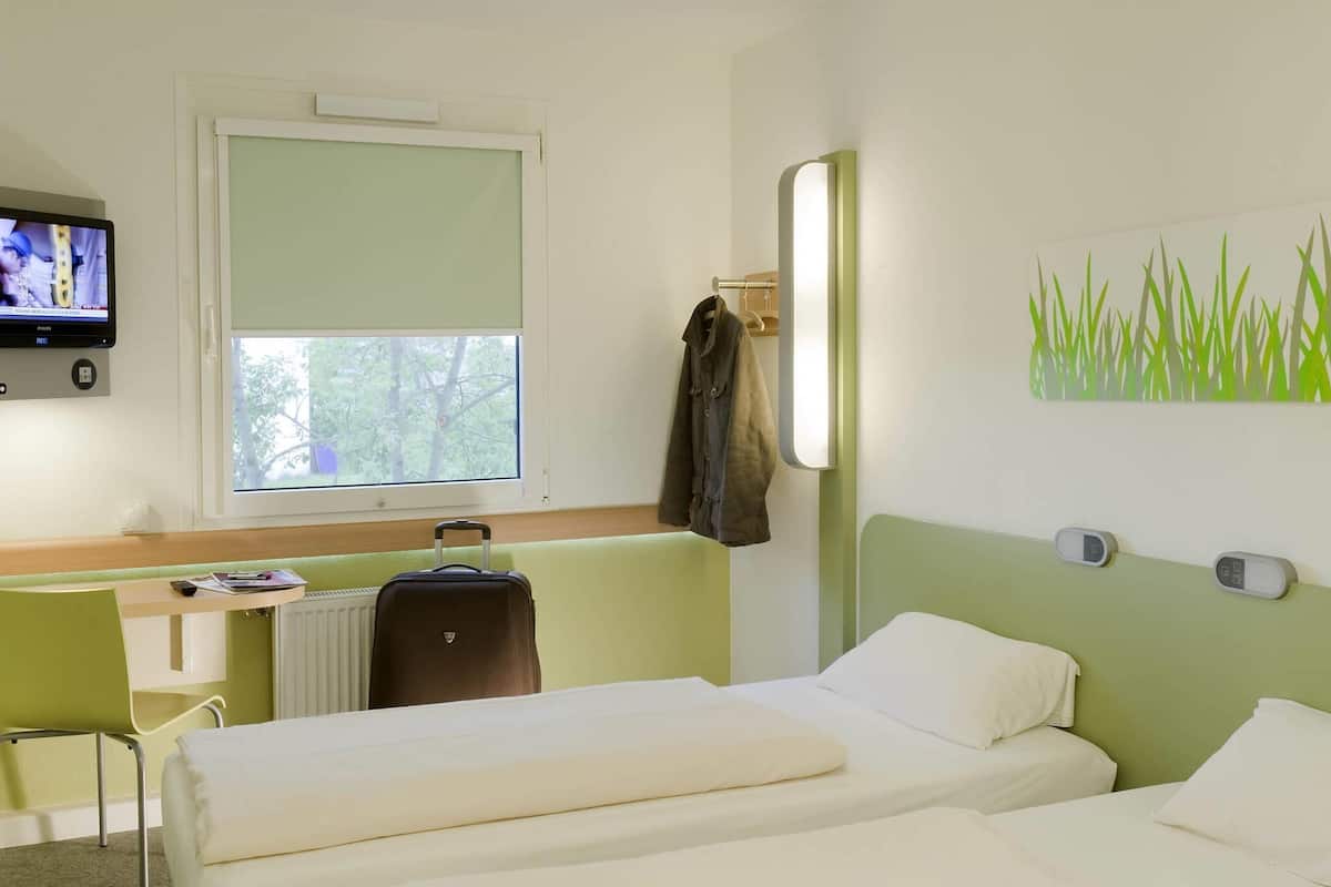 A hotel room with a bed, a chair, a TV, a window, and a coat rack.