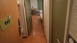 Un pasillo con una puerta de madera que indica '111', un accesorio de iluminación y una vista parcial de una habitación con cama y ventana.
