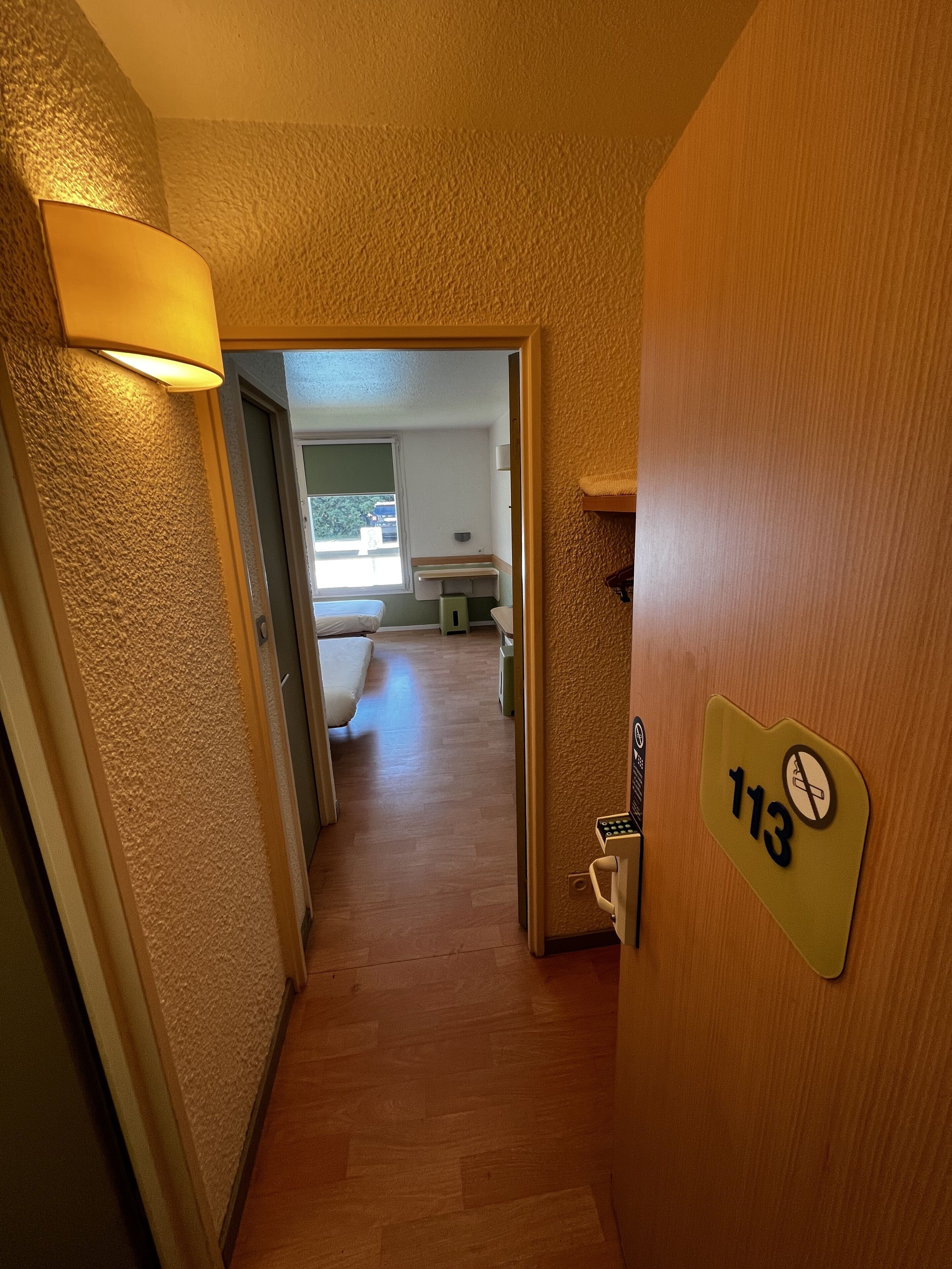 Un pasillo con suelo de madera, una puerta con el número 113 y una lámpara en la pared.