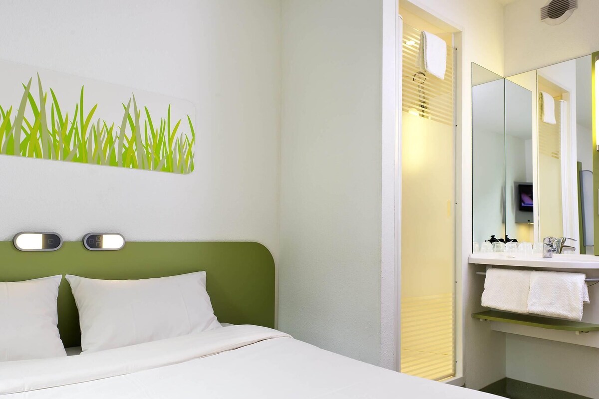 Una habitación de hotel con cama, cabecera verde, luz de pared, espejo y baño con lavamanos y toallero.