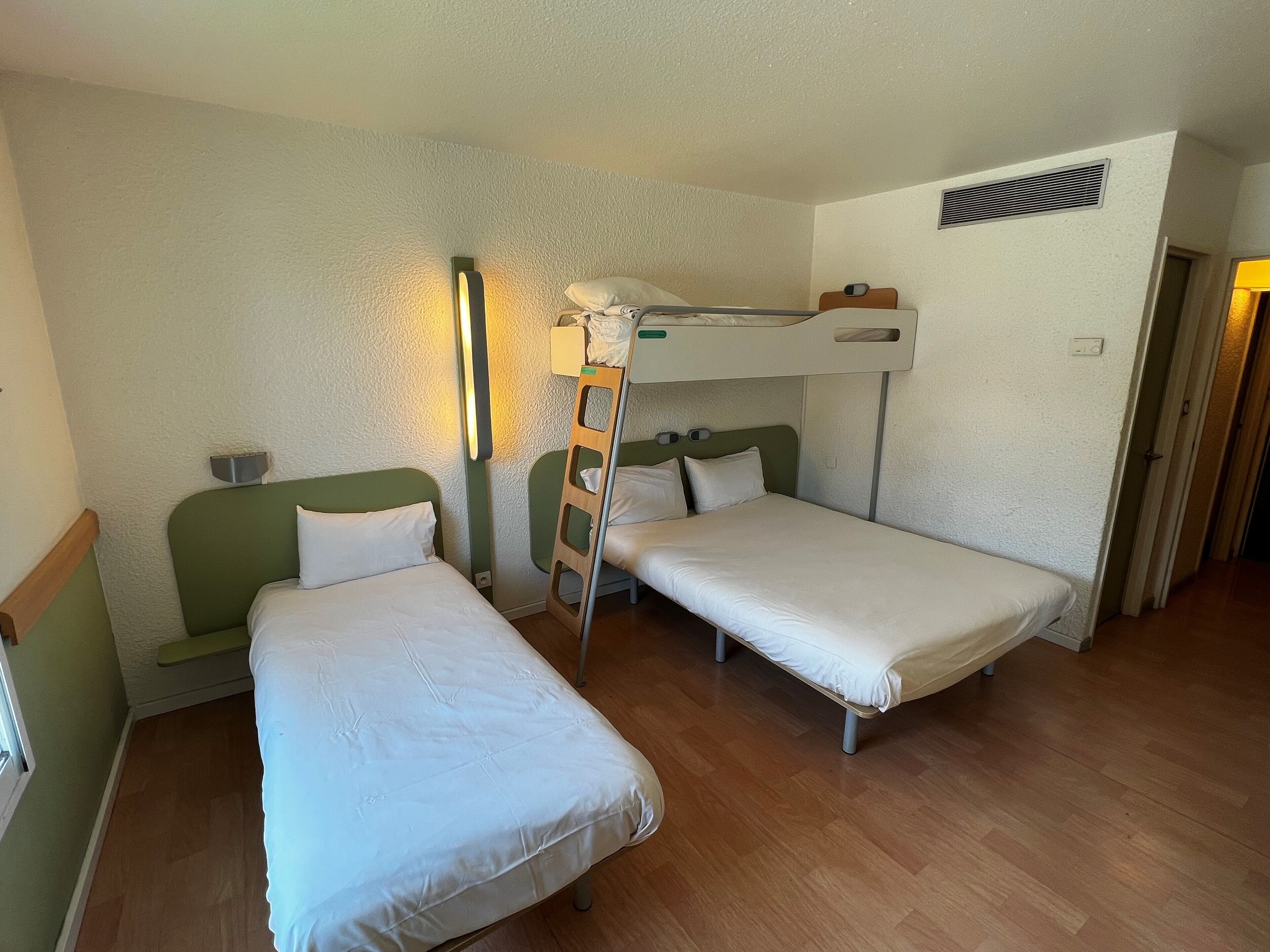 A room with two single beds, a bunk bed, a bedside table with a lamp, and a wooden floor.
