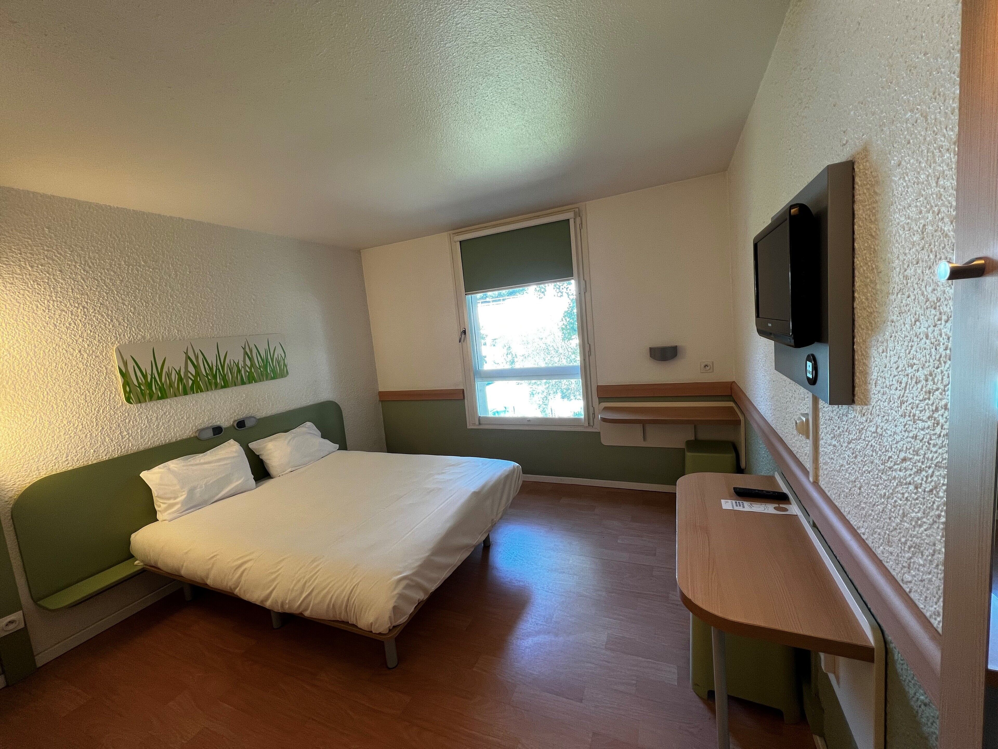 A bedroom with a bed, a desk, a television, and a window.