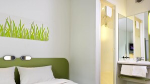 Habitación de hotel con una cama, cabecera verde, luz adosada a la pared, baño con espejo y un perchero.