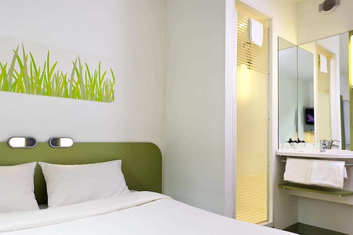 A hotel room with a bed, a green headboard, a wall-mounted light, a bathroom with a mirror, and a towel rack.
