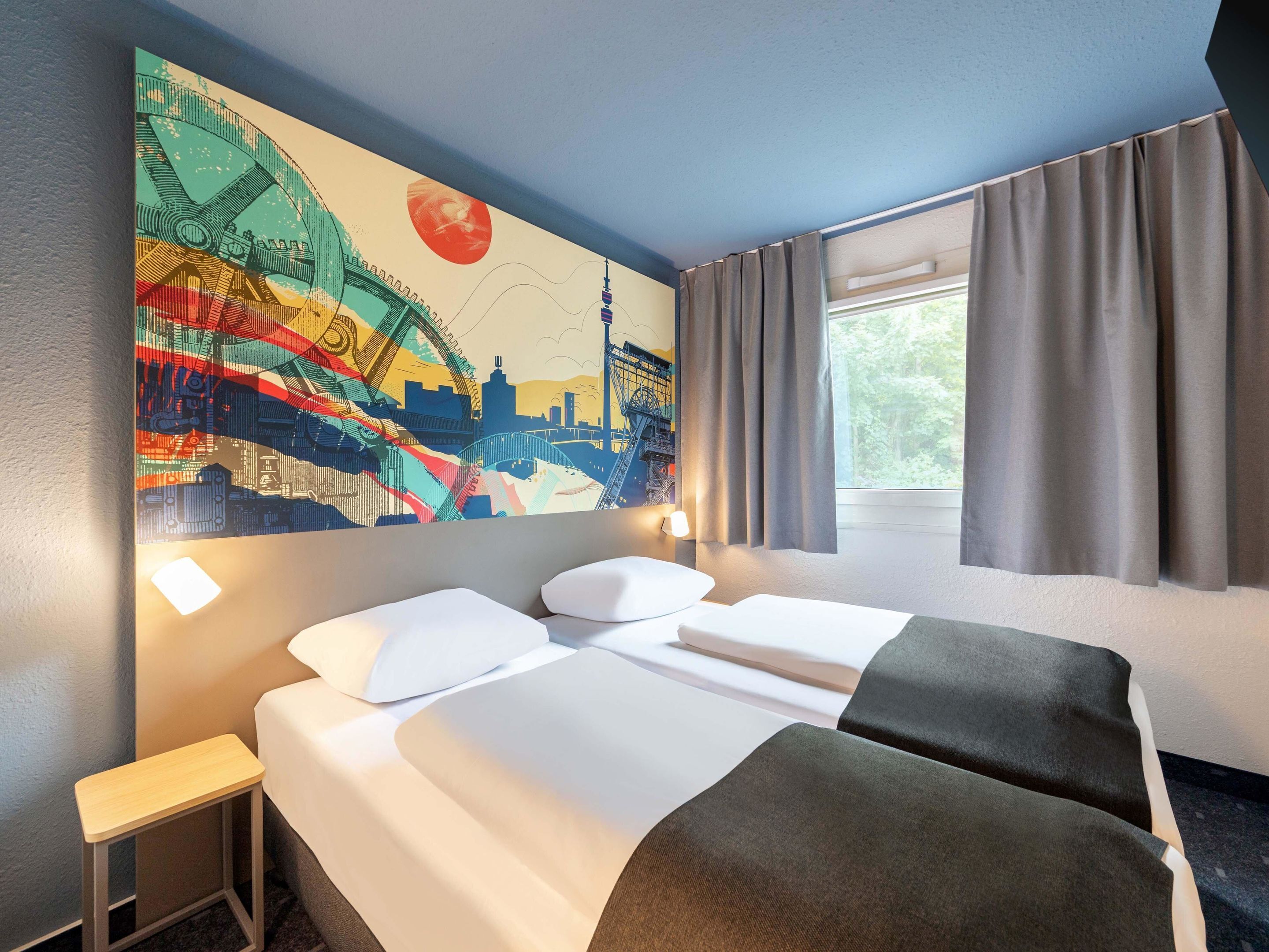 Habitación de hotel con cama, mesita de noche, lámpara y un mural grande con vistas a la ciudad.
