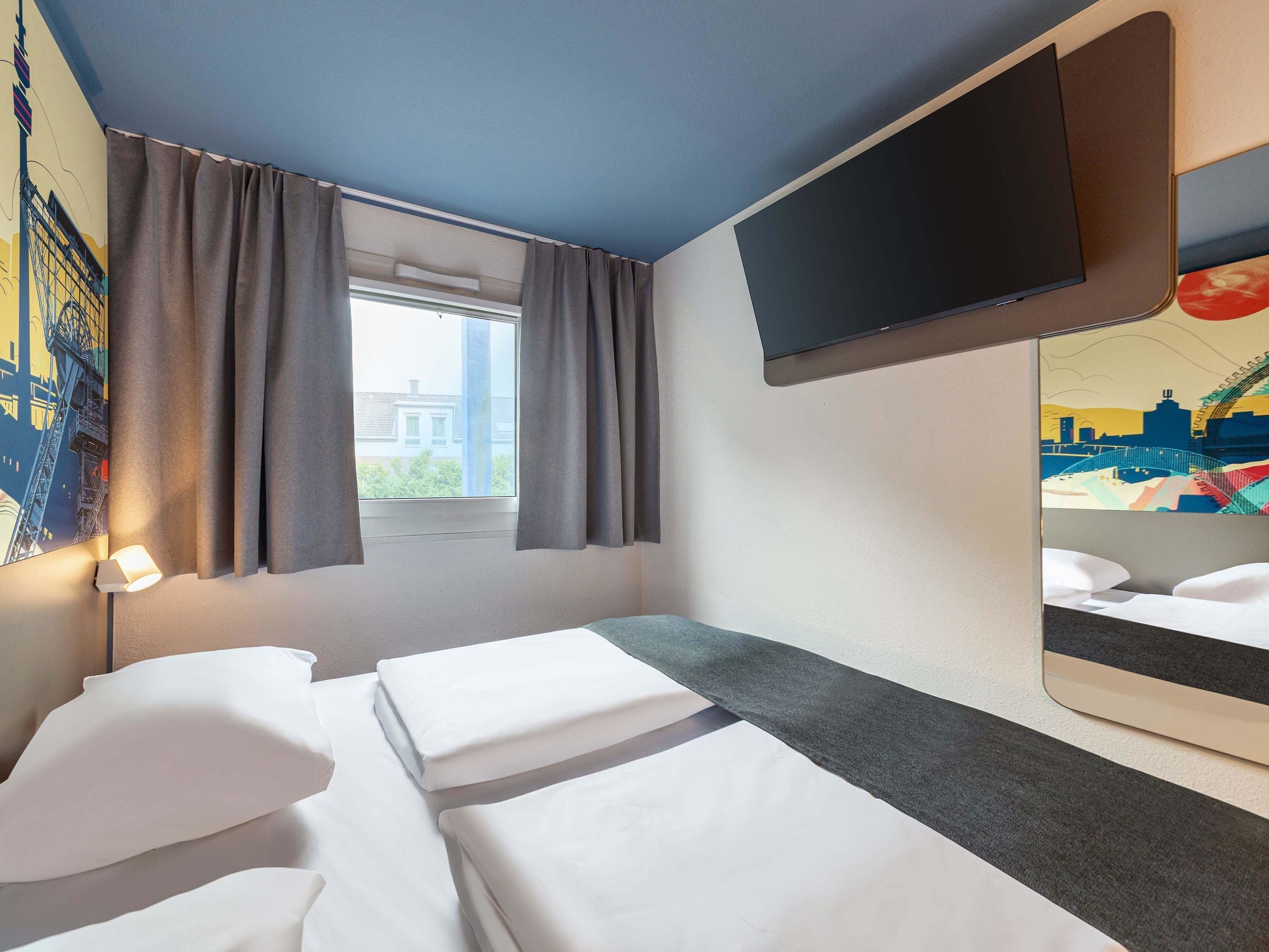 A hotel room with two beds, a flat-screen TV mounted on the wall, a window with curtains, and a cityscape mural on the wall.