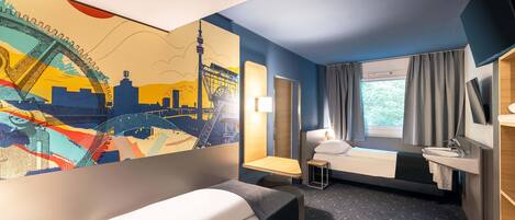 A hotel room with a bed, a small desk, a window with curtains, a wall-mounted TV, and a cityscape mural.