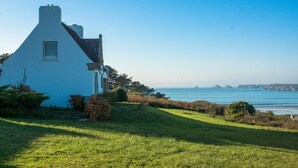 Une maison blanche avec une petite fenêtre et une cheminée, située sur une colline verdoyante qui surplombe un plan d'eau et de lointaines falaises.