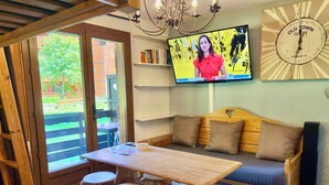 Um quarto aconchegante com uma mesa de madeira, quatro banquinhos, um sofá com almofadas, uma televisão e um relógio na parede.