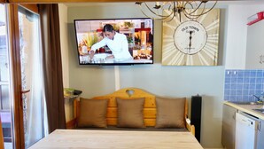 Um quarto aconchegante com uma mesa de jantar de madeira, quatro cadeiras, um sofá com almofadas, uma televisão presa à parede e um relógio sobre o sofá.