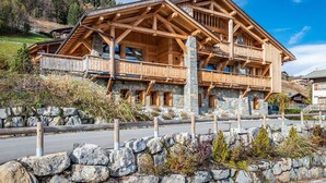 Un chalet en bois à deux étages, doté de fondations en pierre, de balcons et d’un mur de soutènement en pierre à l’avant.