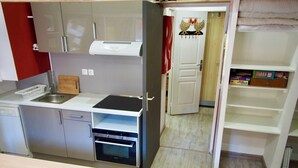 A compact kitchen with a dishwasher, oven, and a small sink. There's a wooden table with a metal bowl and a decorative item. A red chair is visible in the foreground. - Risoul - Studio 08 - Fully equipped - WIFI - 30m² - Foot of ski area (Risoul 1850)