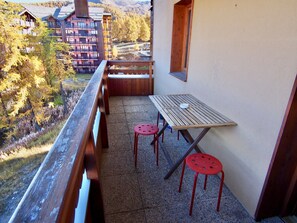 A balcony with a wooden table and two red stools, overlooking a mountainous area with buildings. - Risoul - Studio 407 - Pool/Sauna - WIFI - Great view - Foot of slopes (Risoul)