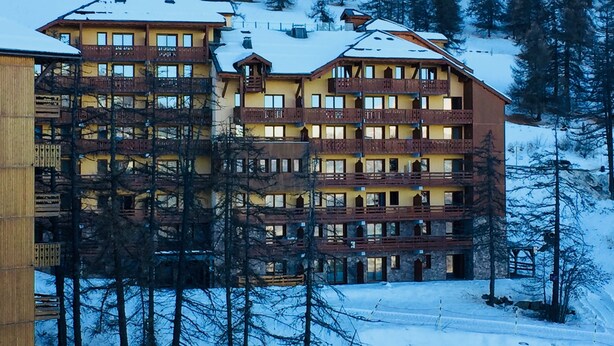 Un hôtel à plusieurs étages, doté de balcons et d’un toit enneigé, entouré d’arbres.