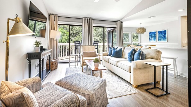 A spacious living room with a sofa, ottoman, and a coffee table. There is a balcony with a chair and a table, and a ceiling fan. - Pool View, Open Plan, Pet Friendly & No Carpeting! (Hilton Head Island)