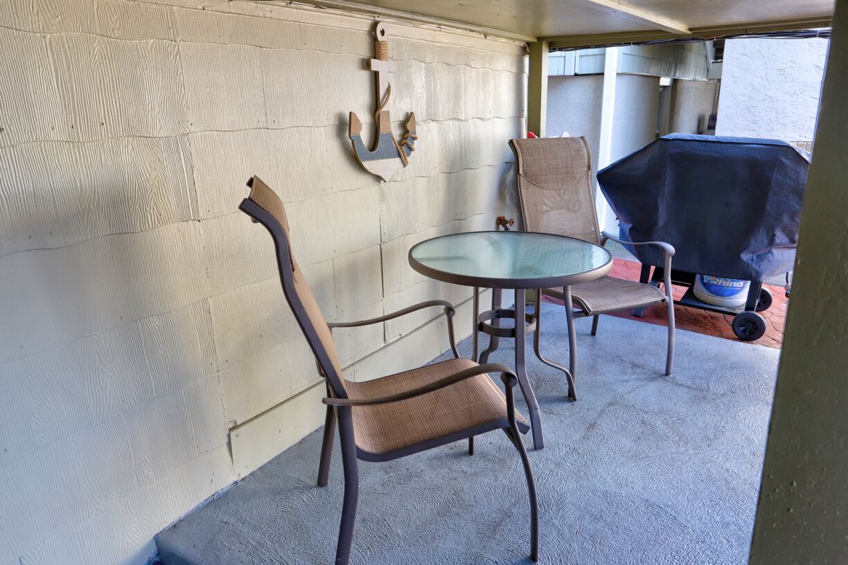 Un patio couvert comprenant une table ronde en verre et deux chaises, un barbecue et une décoration murale en forme d’ancre.
