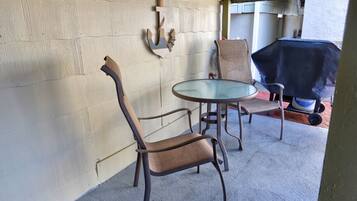 Un patio couvert comprenant une table ronde en verre et deux chaises, un barbecue et une décoration murale en forme d’ancre.