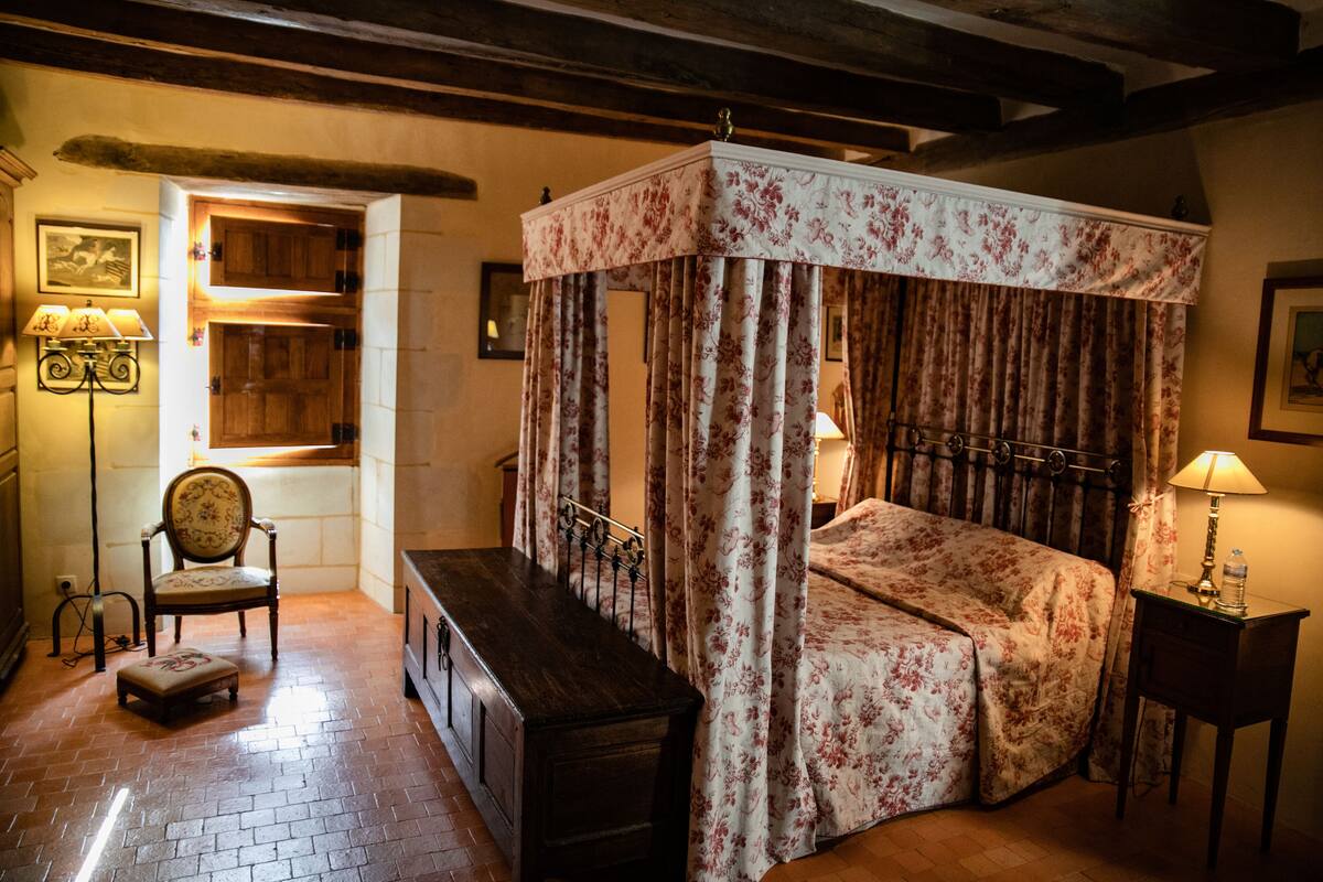 Un lit à baldaquin avec des rideaux à motifs floraux, un banc en bois, une chaise et une table de chevet avec une lampe.