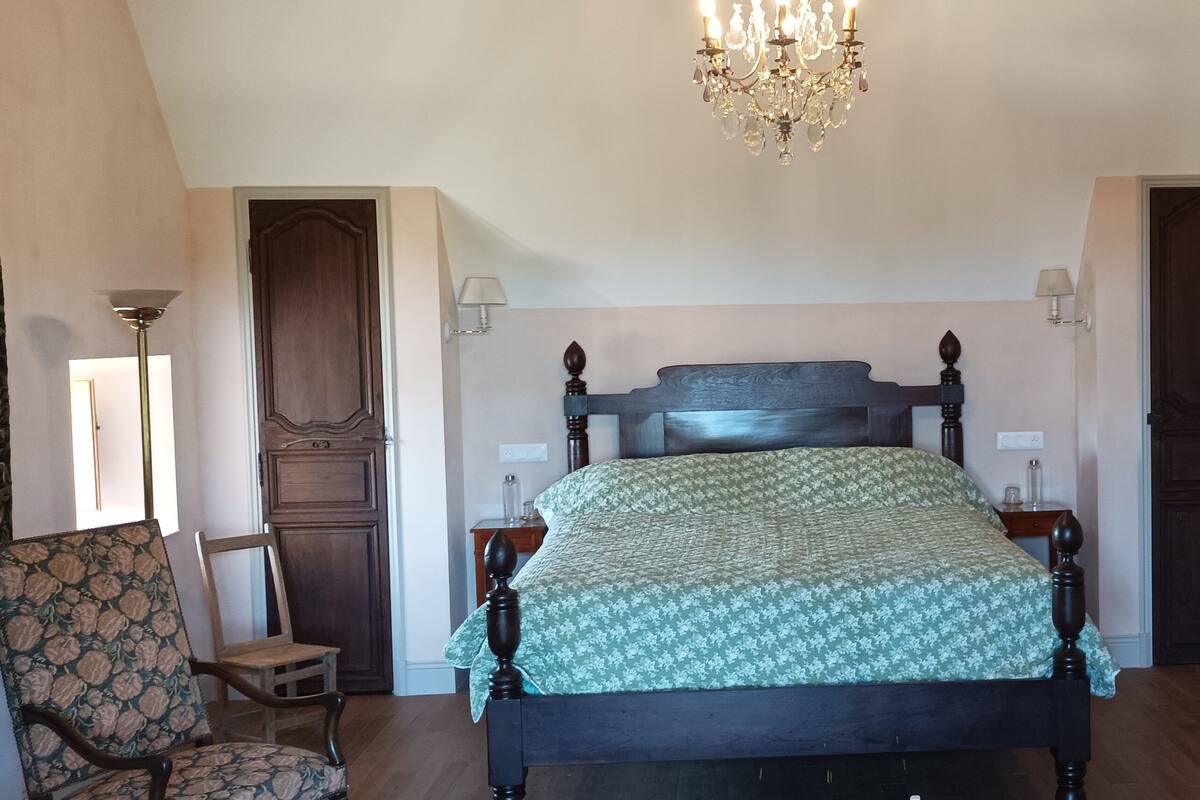 Une chambre à coucher avec un grand lit, deux tables de chevet, une chaise, un petit tabouret, un lustre et une porte.