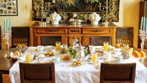 Une table à manger dressée avec des aliments pour le petit-déjeuner, une tapisserie à motifs floraux et une horloge ancienne accrochée au mur.