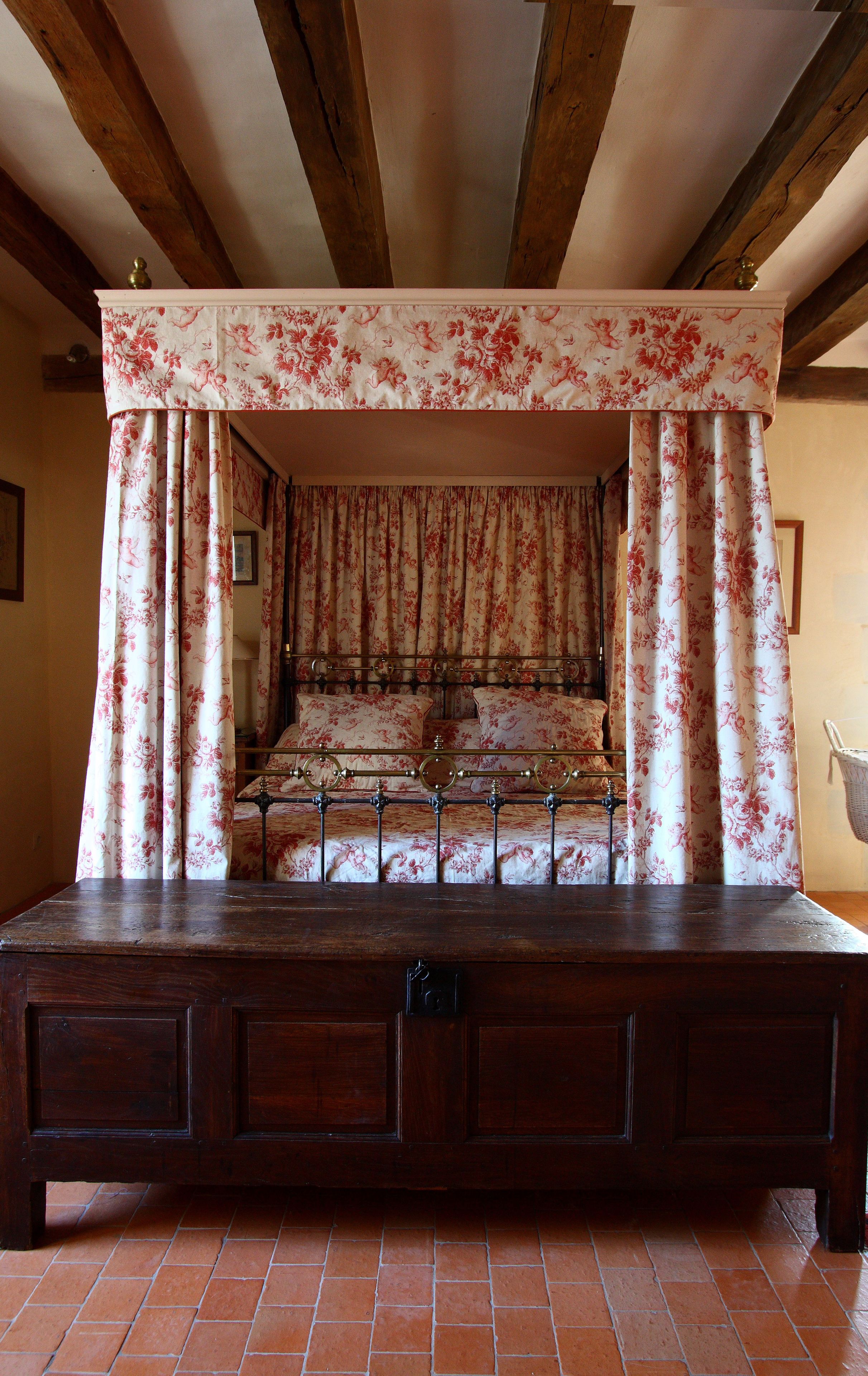 Un lit à baldaquin à quatre poteaux, agrémenté de rideaux à motifs floraux, un banc en bois devant, et des poutres apparentes en bois au plafond.