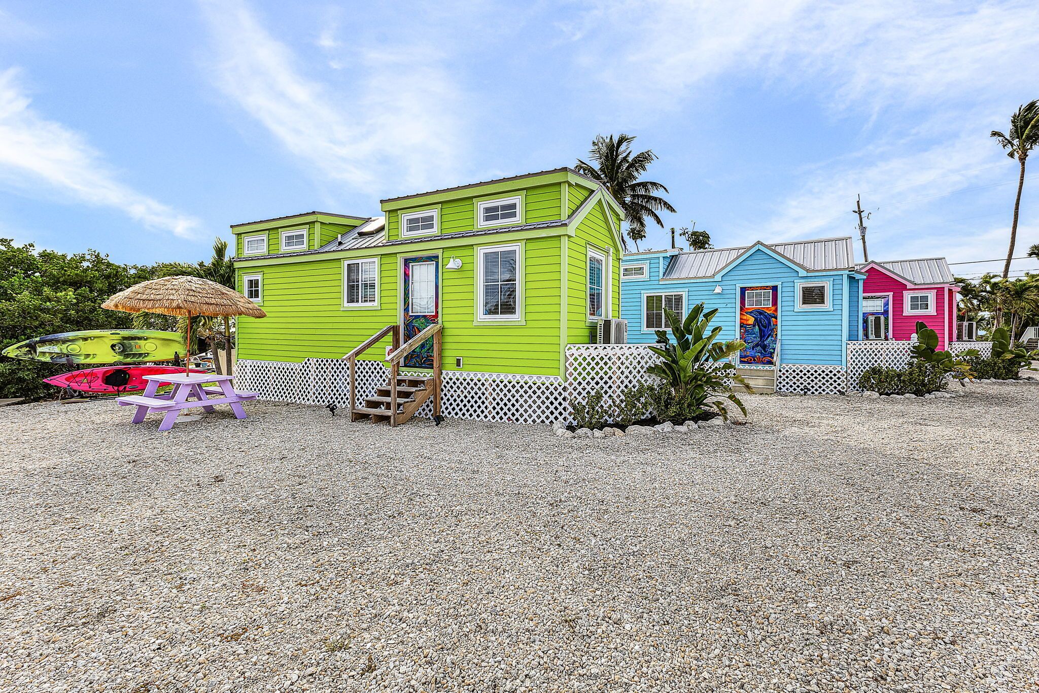 Een kleurrijk pand aan het strand met een groen huis, een blauw huis en een roze huis. Er is een houten trap die naar het groene huis leidt, en een rieten parasol en een paarse tafel in de buurt.