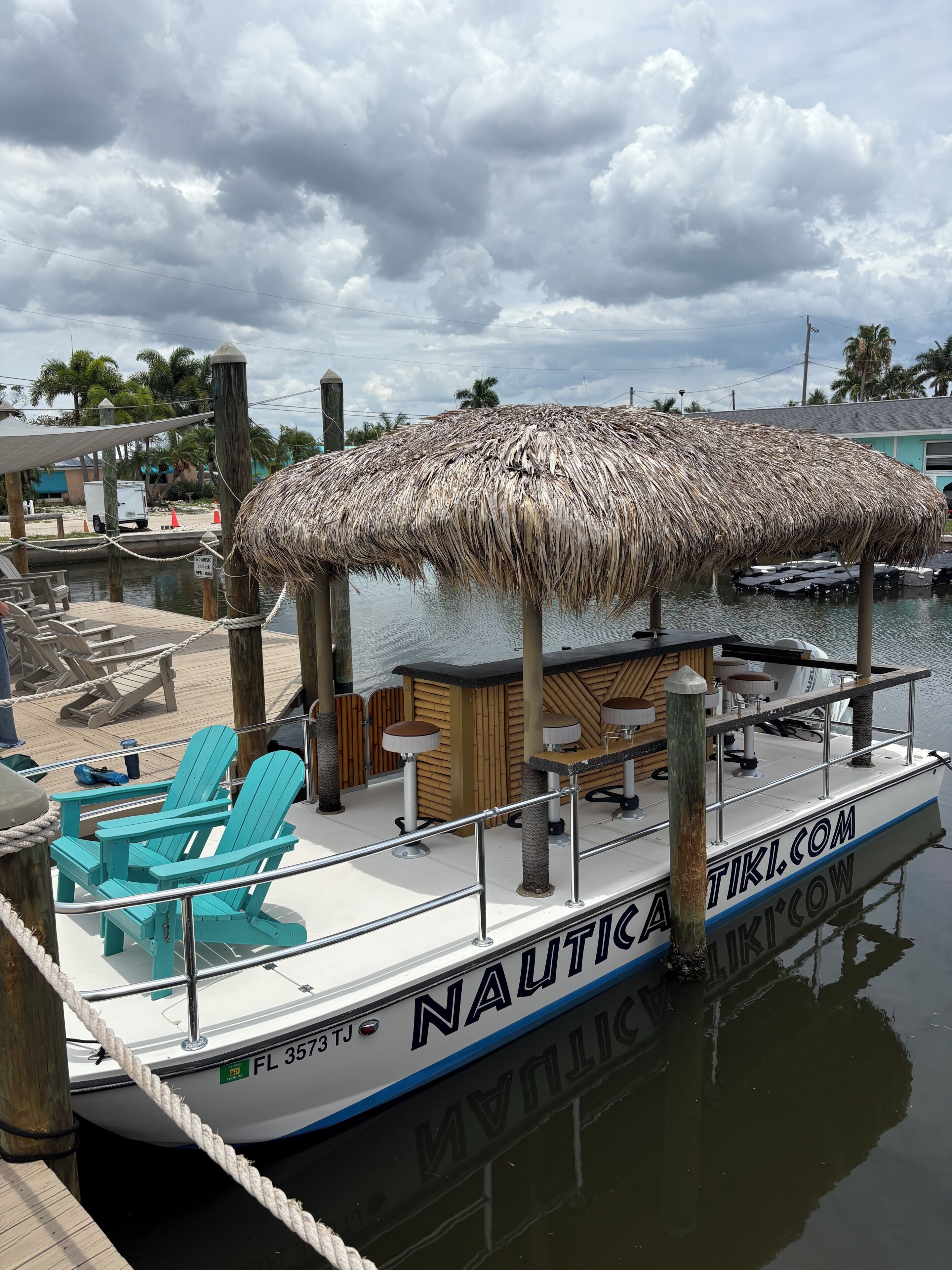 A boat with a thatched roof, named 'NAUTICA TIKI.COM,' docked at a marina with lounge chairs and a wooden deck.