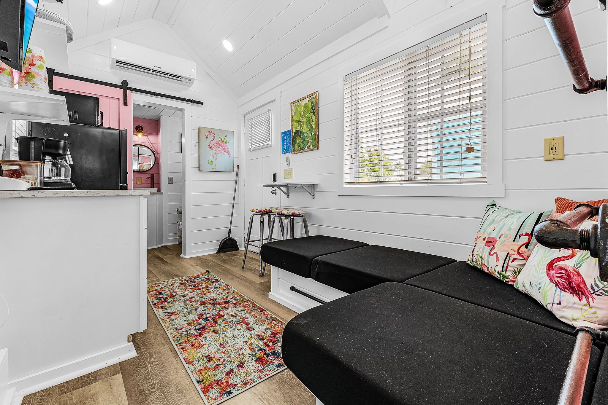 A compact living space with a kitchenette, a black sofa, a colorful rug, and a window with blinds.