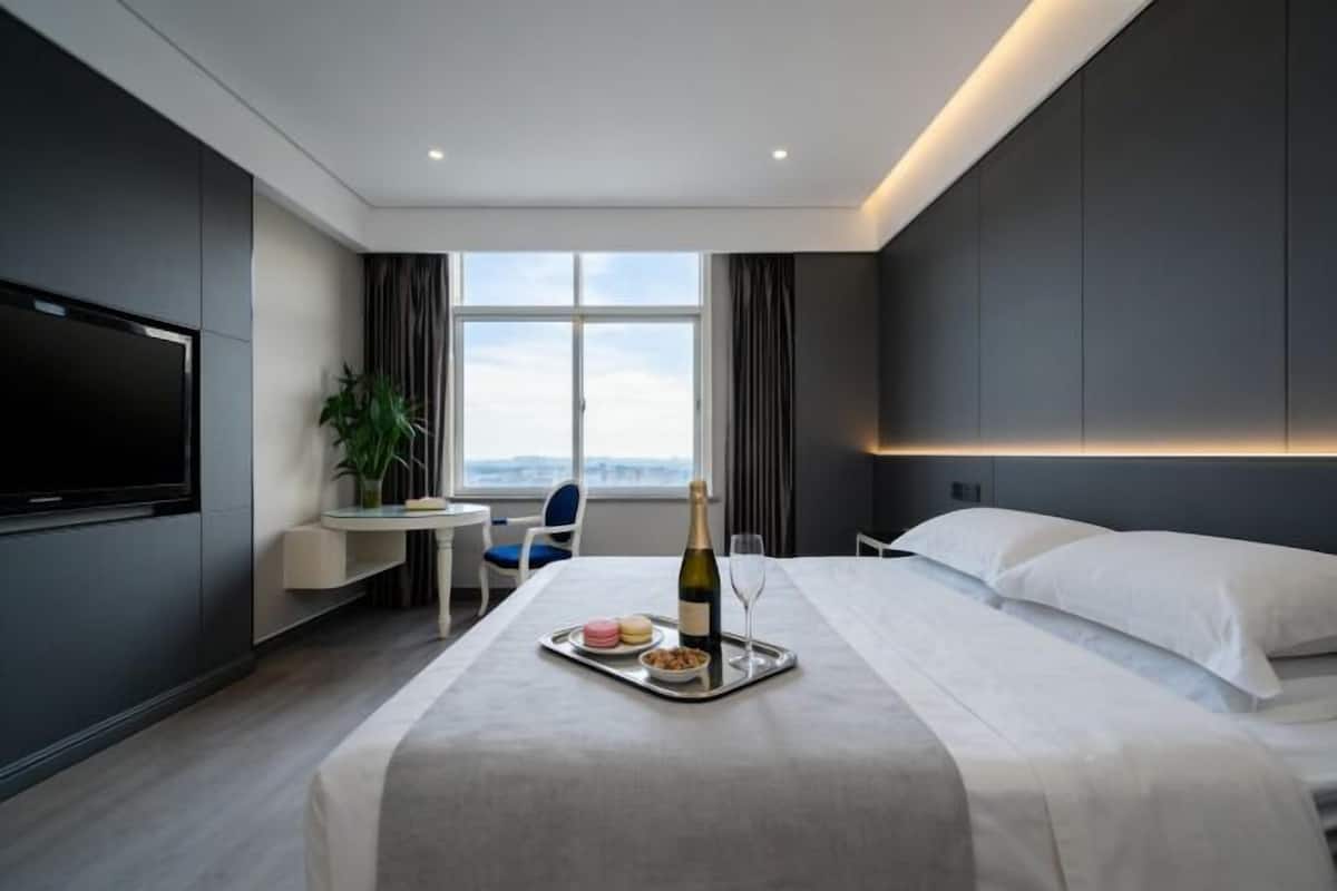 A modern hotel room with a bed, a desk with a chair, a TV, a large window, and a bottle of champagne on a tray.