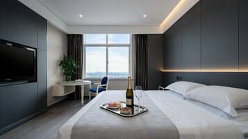 A modern hotel room with a bed, a desk with a chair, a TV, a large window, and a bottle of champagne on a tray.