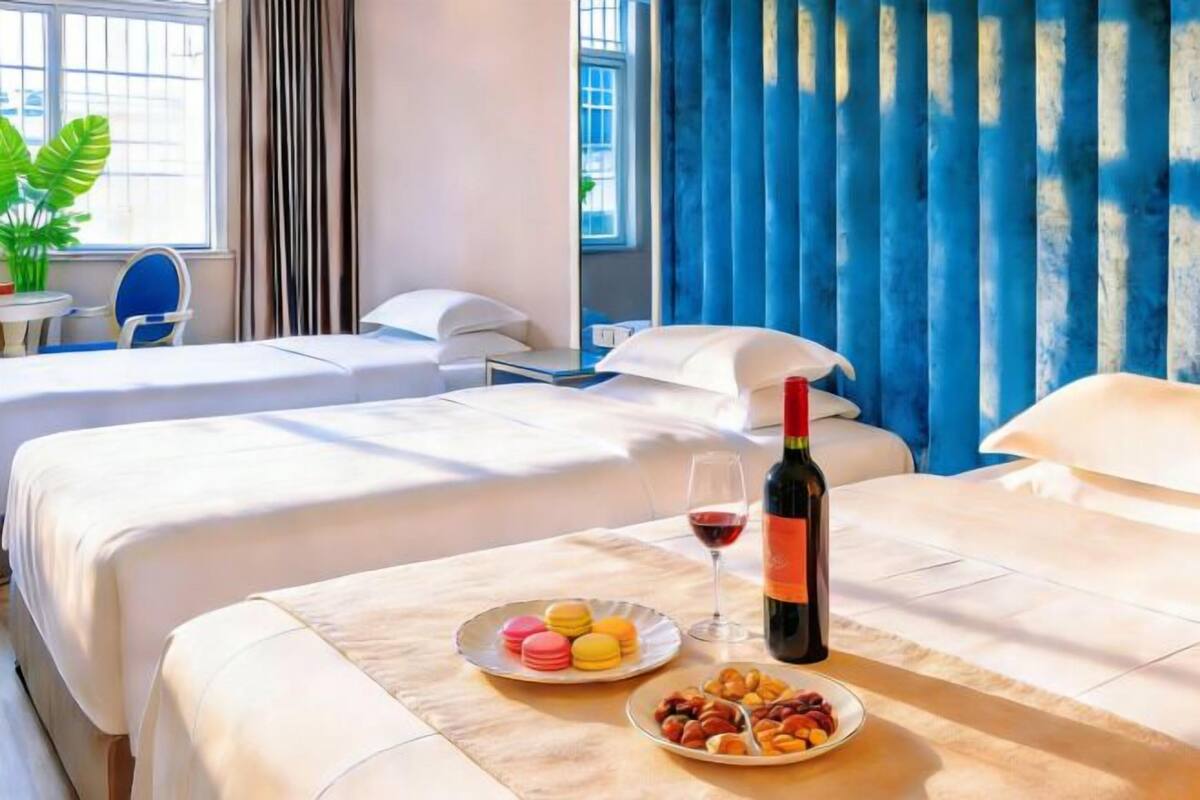 A hotel room with two beds, a dining table with a bottle of wine, a glass of wine, and plates of macarons and nuts.