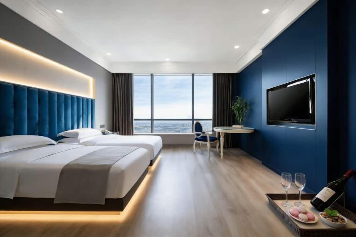 A modern hotel room with two beds, a TV, a dining area, and a view of the city.