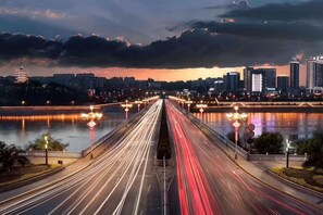 Egy városi táj, amelyen folyó, híd és hosszú expozíciós idővel készült, mozgó járműveket ábrázoló felvétel látható.