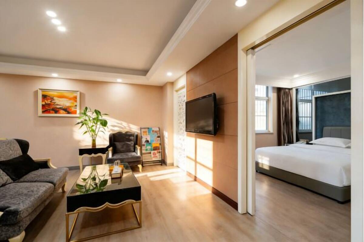 A modern hotel room with a bed, a sofa, a coffee table, a TV, and a painting on the wall.