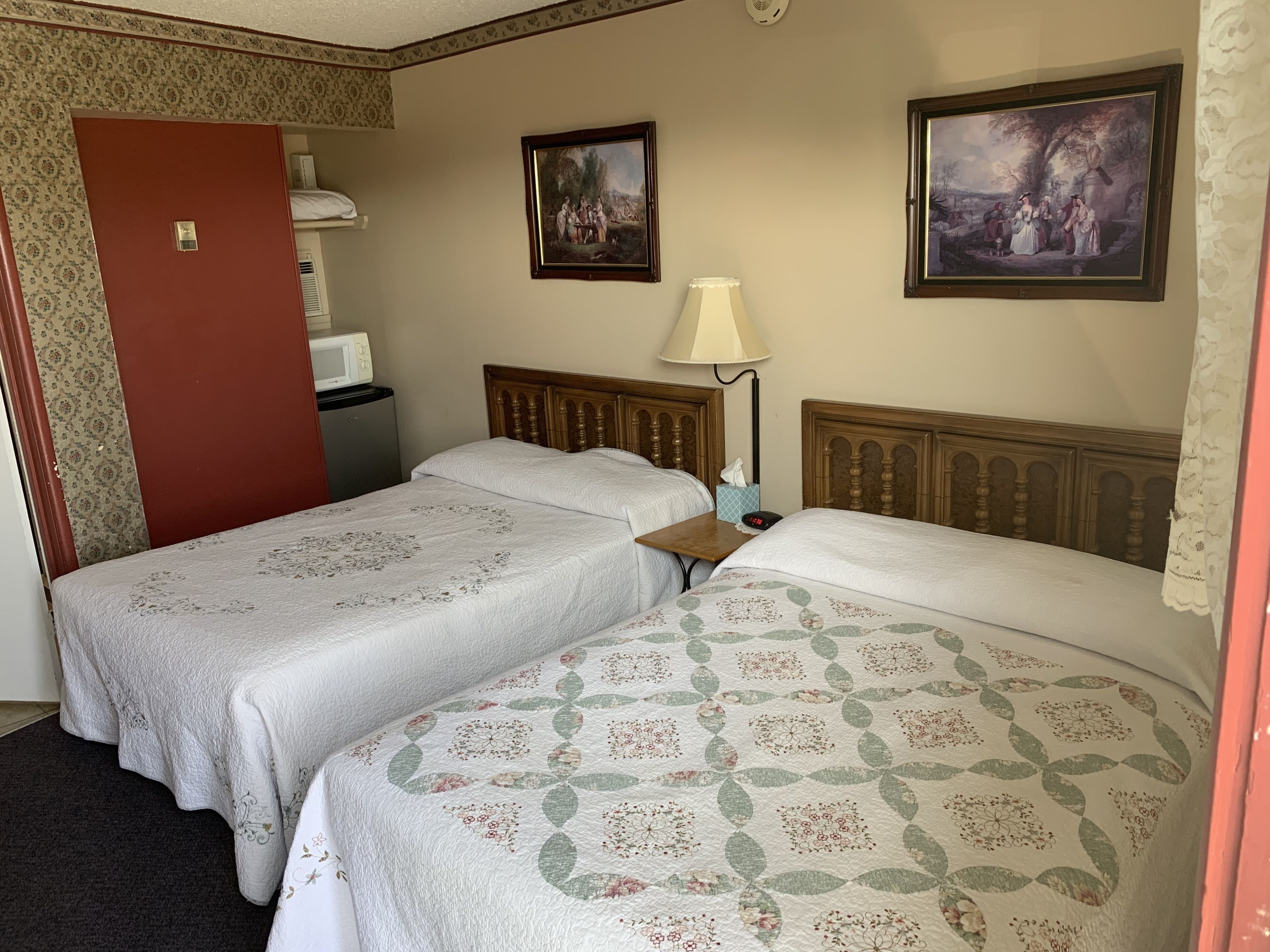 A hotel room with two beds, a bedside table, a lamp, a clock, and two framed pictures on the wall.