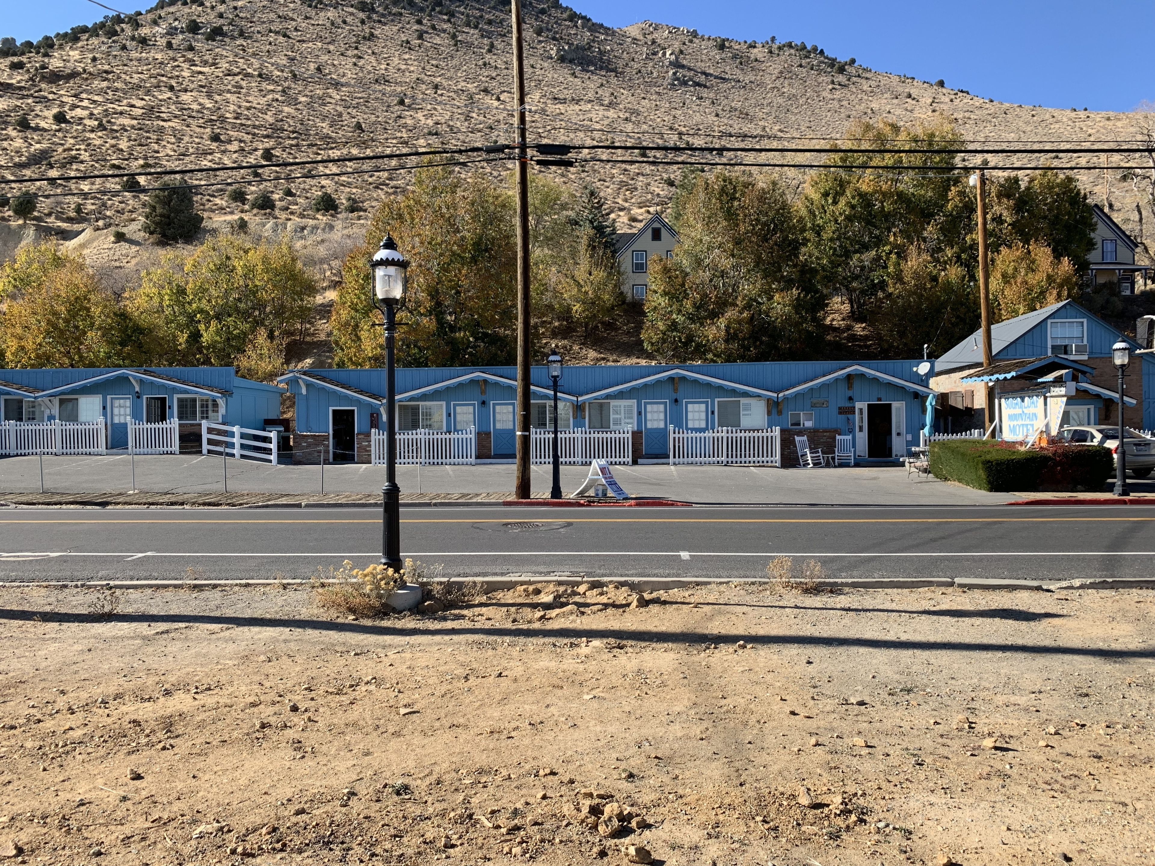 A row of blue motel buildings with white picket fences, a parking lot, and a hilly backdrop.