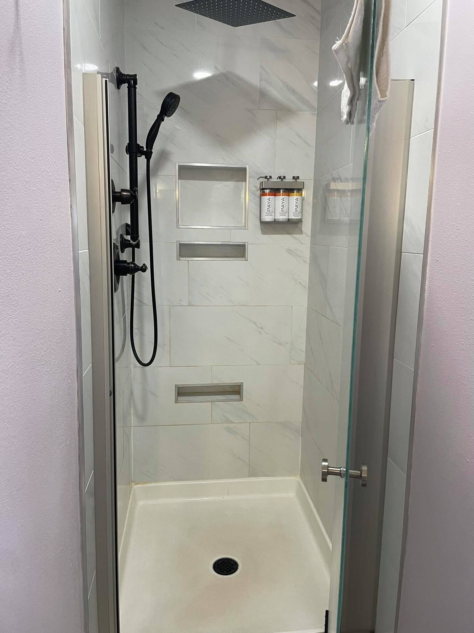 Une douche à l’italienne avec une porte en verre, une douchette à main et un distributeur de savon fixé au mur.
