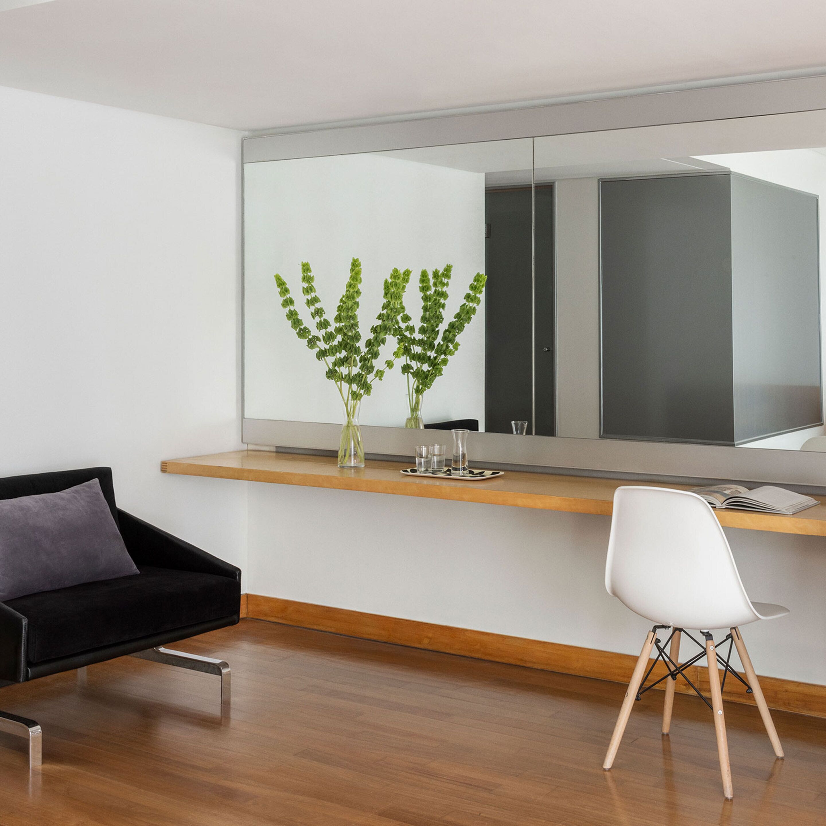 A modern room with a wooden floor, a black sofa, a white chair, and a large mirror above a wooden shelf with a vase of green plants.