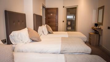 A hotel room with three beds, a wooden door, a bathroom visible through an open door, and a small wooden side table with a mirror.