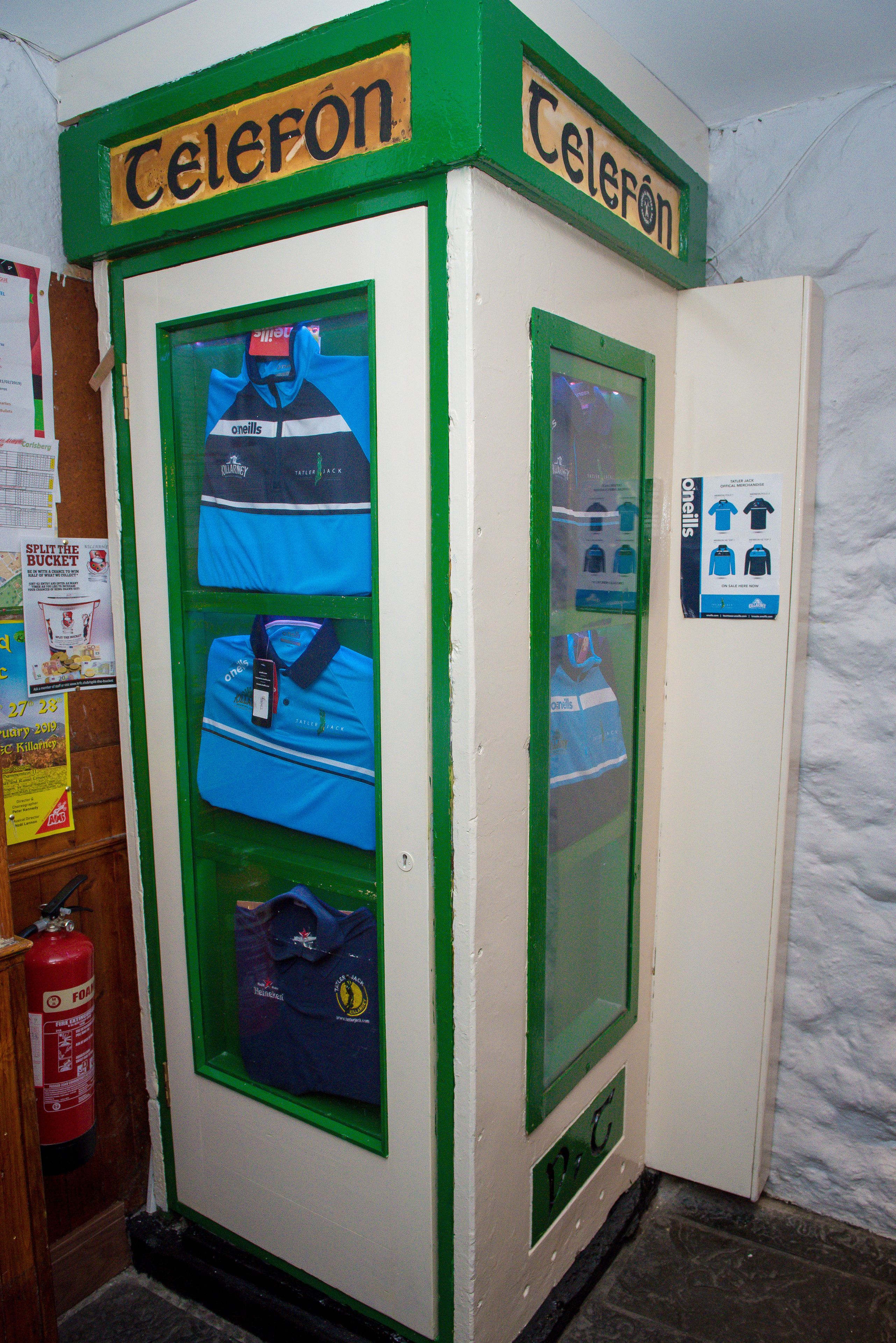 A telephone booth with a green frame and a sign that reads "Telefon". Inside, there are blue bags and a blue shirt displayed.