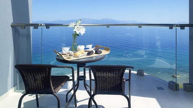 Un balcon avec une table dressée pour le petit-déjeuner, deux chaises en osier et une vue dégagée sur la mer et les montagnes lointaines.